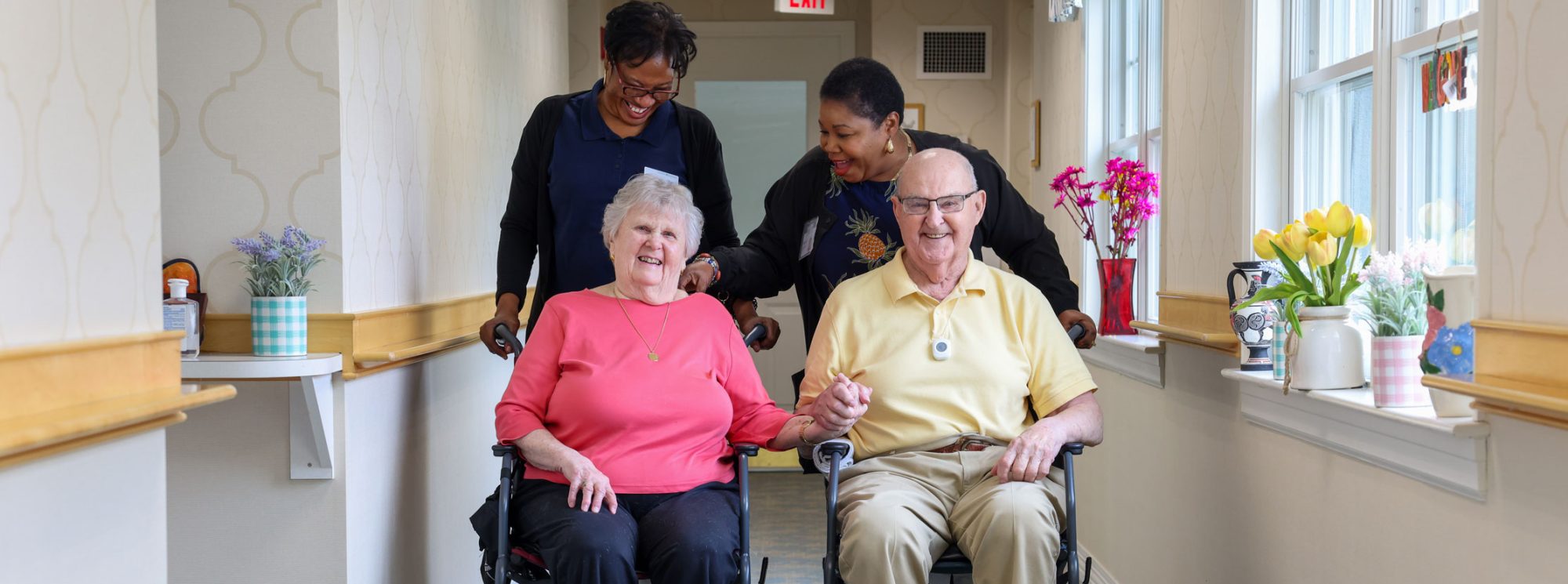 A couple happily posing for the camera in their wheelchairs, with CNAs happily standing behind them.